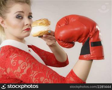 Dieting, weight loss concept. Funny blonde woman holding yummy choux puff cake with whipped cream and boxing glove, fighting off bad food. On grey. Woman fighting off bad food, boxing cream puff cake
