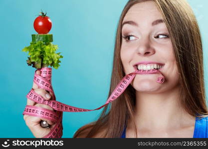 Dieting weight loss concept. Sporty girl fitness woman holding fork with fresh mixed vegetables and measuring tape on blue background