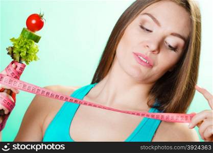 Dieting weight loss concept. Sporty girl fitness woman holding fork with fresh mixed vegetables and measuring tape on green-blue background.