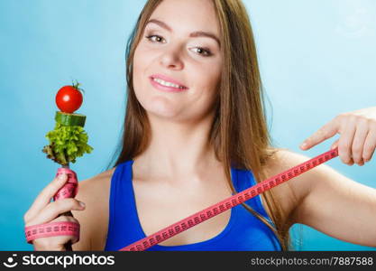 Dieting weight loss concept. Sporty girl fitness woman holding fork with fresh mixed vegetables and measuring tape on blue background.