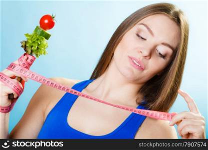 Dieting weight loss concept. Sporty girl fitness woman holding fork with fresh mixed vegetables and measuring tape on blue background.