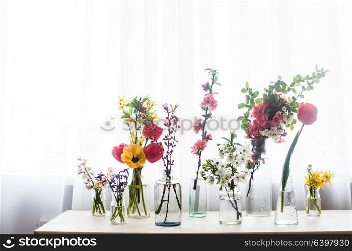 Different beautiful flowers in jars with water on the table near the window. Group of different flowers on the table