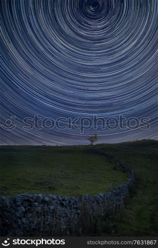 Digital composite image of star trails around Polaris with Beautiful Autumn Fall landscape countryside image of lone tree and stone wall
