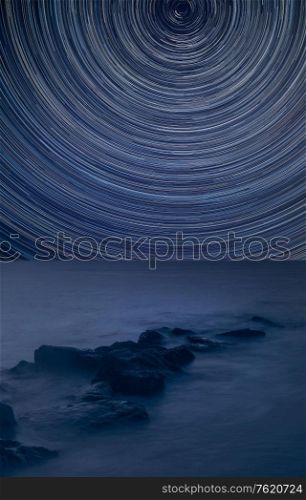 Digital composite image of star trails around Polaris with Peaceful cross processed landscape image of calm sea over rocks