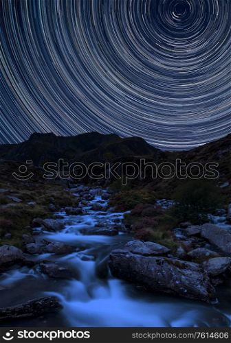 Digital composite image of star trails around Polaris with Stunning vibrant Landscape image of river flowing down mountain range