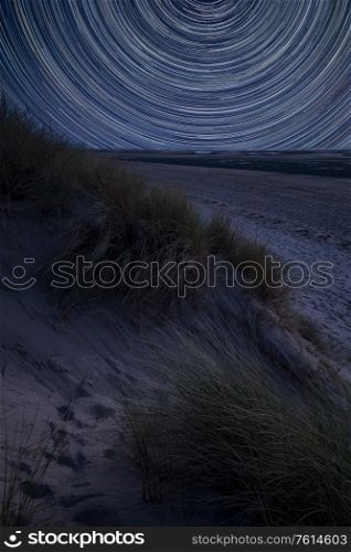 Digital composite image of star trails around Polaris with Summer landscape over grassy sand dunes on beach