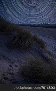 Digital composite image of star trails around Polaris with Summer landscape over grassy sand dunes on beach