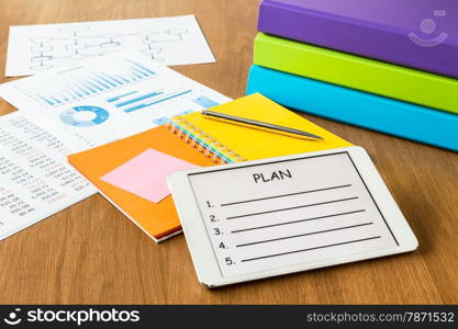 Digital tablet pc showing blank form of project planning on a display screen, colorful diary book, binders and documents on workspace