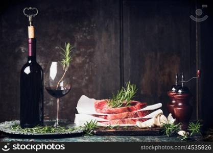 Dinner preparation with raw steaks, Bottle and glass of red wine ,rosemary and salt and pepper shakers on kitchen table at dark wooden background, side view,place for text