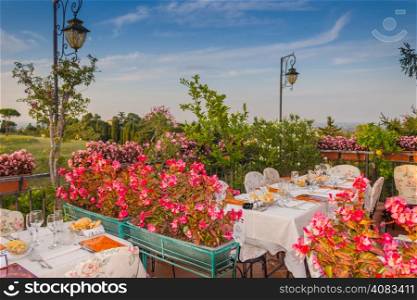 Dinner tables in elegant style Italian outdoor restaurant on iron grating parapet on the hills of Dozza (Bo) in Italy in a summer sunny day: real red, yellow and pink flowers with green leaves in green pots all around. Pink napkins and tablecloth, classy set of silver cutlery and crystal glasses