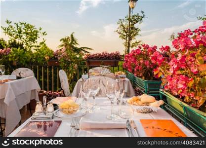 Dinner tables in elegant style Italian outdoor restaurant on iron grating parapet on the hills of Dozza (Bo) in Italy in a summer sunny day: real red, yellow and pink flowers with green leaves in green pots all around. Pink napkins and tablecloth, classy set of silver cutlery and crystal glasses