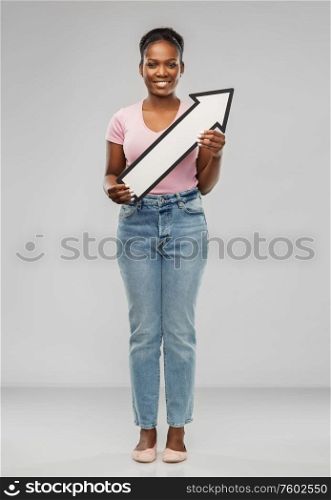 direction and people concept - happy african american young woman holding big white thick arrow showing to north east over grey background. african woman with arrow showing north east