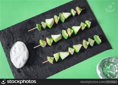 Directly above view of a black platter with green fruits skewers, yogurt dip and a glass of water. Diet food. Detoxification meal. Healthy eating.