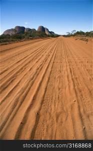 Dirt road with tire tracks in Uluru Kata Tjuta National Park, Australia with Mount Olga in the distance.