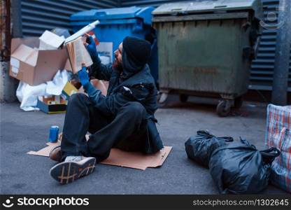 Dirty drunk beggar lies in garbage at the trashcan on city street. Poverty is a social problem, homelessness and loneliness, alcoholism and drunk addiction, urban lonely. Dirty drunk beggar lies in garbage at the trashcan