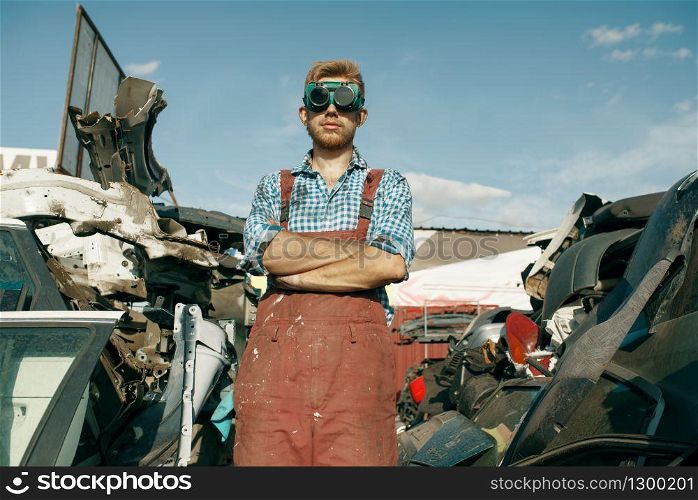 Dirty male repairman in welding glasses on car junkyard. Auto scrap, vehicle junk, automobile garbage. Abandoned, damaged and crushed transport, scrapyard