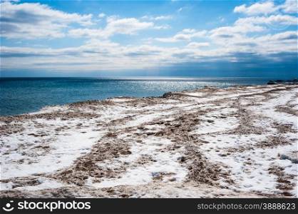 Dirty white and brown ice and snow coastline against bright partly cloudy blue sky and dark blue water horizon.