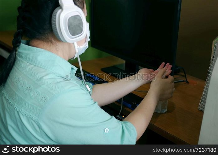Disability blind person with headphone wearing face mask applying alcohol gel hand sanitizer on hands before using computer with braille display amid Coronavirus (COVID-19) pandemic.