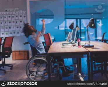 Disabled businessman in wheelchair working overtime alone at his desk on a desktop computer in an open space modern coworking office late at night with city lights glowing in the background. . businessman working overtime alone at his desk in an office late at night 