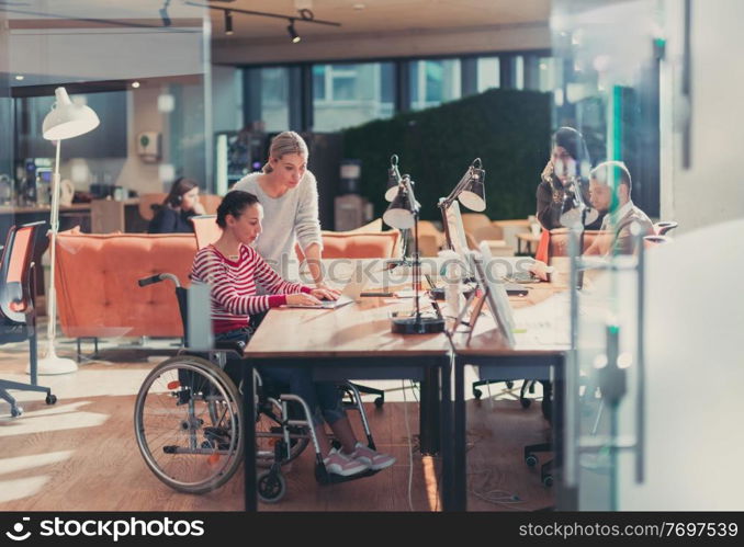 Disabled businesswoman in a wheelchair working in a creative office. Business team in modern coworking office space. Colleagues working in the background at late night. Inclusion and handicap concept.. Disabled businesswoman in a wheelchair in modern coworking office space. Colleagues in background. Disability and handicap concept. Selective focus