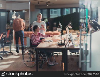 Disabled businesswoman in a wheelchair working in a creative office. Business team in modern coworking office space. Colleagues working in the background at late night. Inclusion and handicap concept.. Disabled businesswoman in a wheelchair in modern coworking office space. Colleagues in background. Disability and handicap concept. Selective focus
