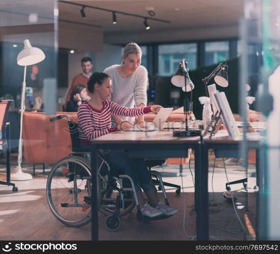 Disabled businesswoman in a wheelchair working in a creative office. Business team in modern coworking office space. Colleagues working in the background at late night. Inclusion and handicap concept.. Disabled businesswoman in a wheelchair in modern coworking office space. Colleagues in background. Disability and handicap concept. Selective focus