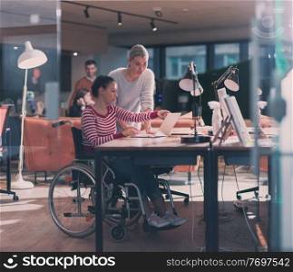 Disabled businesswoman in a wheelchair working in a creative office. Business team in modern coworking office space. Colleagues working in the background at late night. Inclusion and handicap concept.. Disabled businesswoman in a wheelchair in modern coworking office space. Colleagues in background. Disability and handicap concept. Selective focus