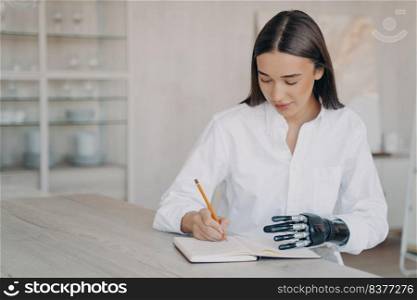 Disabled european girl is taking notes at the desk and doing homework. Happy handicapped young woman with futuristic cyber arm is studying remotely in college. Concept of equal opportunities.. Disabled european girl with cyber arm is taking notes at the desk and doing homework.
