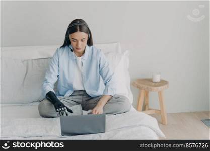 Disabled european woman with artificial arm working on laptop in bedroom. Freelancer has online meeting sitting on her bed using headset and microphone. Handicapped girl is remote assistant.. Disabled european woman with artificial arm is working on laptop in bedroom sitting on her bed.