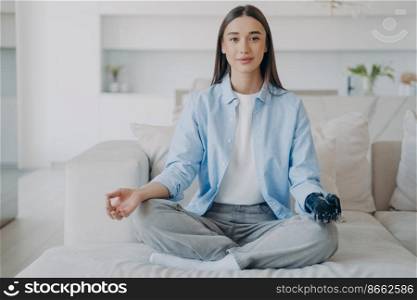 Disabled female does yoga, sitting in lotus pose on couch, folded fingers in mudra gesture. Calm girl with bionic prosthetic arm does breathing exercises on sofa at home. Disability and stress relief.. Disabled girl with bionic prosthetic arm does yoga, sitting in lotus pose. Disability, stress relief