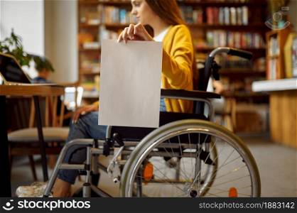 Disabled female student in wheelchair holds empty paper sheet, disability, bookshelf and university library interior on background. Handicapped woman in college, paralyzed people get knowledge. Disabled student in wheelchair holds paper sheet