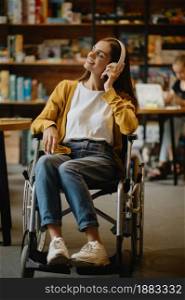 Disabled female student in wheelchair listen to music in headphones, disability, bookshelf and library interior on background. Handicapped woman studying in college, paralyzed people get knowledge. Disabled student listen to music in headphones