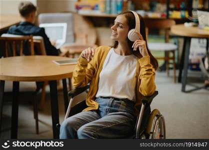 Disabled female student in wheelchair listen to music in headphones, disability, bookshelf and library interior on background. Handicapped woman studying in college, paralyzed people get knowledge. Disabled student listen to music in headphones