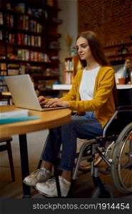 Disabled female student in wheelchair using laptop, disability, bookshelf and university library interior on background. Handicapped young woman studying in college, paralyzed people get knowledge. Disabled female student in wheelchair using laptop