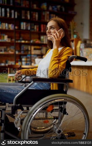 Disabled female student in wheelchair using phone, disability, bookshelf and university library interior on background. Handicapped young woman studying in college, paralyzed people get knowledge. Disabled female student in wheelchair using phone