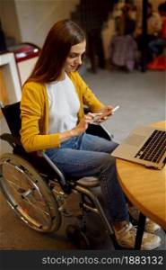 Disabled female student in wheelchair using phone, disability, bookshelf and university library interior on background. Handicapped young woman studying in college, paralyzed people get knowledge. Disabled female student in wheelchair using phone