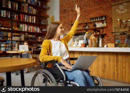 Disabled female student in wheelchair with raised hand, disability, bookshelf and university library interior on background. Handicapped woman studying in college, paralyzed people get knowledge. Disabled student in wheelchair with raised hand