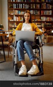Disabled female student in wheelchair works on laptop, disability, bookshelf and university library interior on background. Handicapped young woman studying in college, paralyzed people get knowledge. Disabled student in wheelchair works on laptop