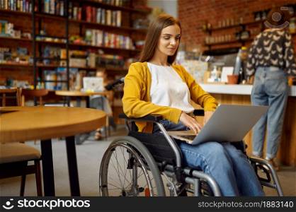Disabled female student in wheelchair works on laptop, disability, bookshelf and university library interior on background. Handicapped young woman studying in college, paralyzed people get knowledge. Disabled student in wheelchair works on laptop