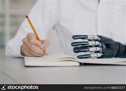 Disabled female writing with pencil in notebook, close-up. Woman with disability notes in notepad, makes to-do list, using bionic prosthetic arm, artificial robotic limb. Medical high tech concept.. Disabled female writing with pencil in notebook, using bionic prosthetic arm, closeup. Medical tech