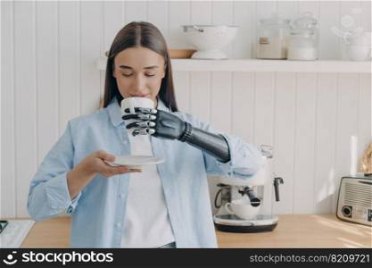 Disabled girl drinking coffee or tea holding mug by high tech bionic prosthetic arm in cozy kitchen. Young woman with disability enjoying morning routine at home using artificial hand.. Disabled girl drinking coffee or tea holding mug using high tech bionic prosthetic arm in kitchen