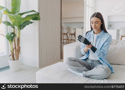 Disabled girl gets rehabilitation, learning to install her bionic prosthetic arm, sitting on couch in cozy modern living room. Young woman with disability training to use high tech artificial limb.. Girl learning to use bionic prosthetic arm in modern living room. Rehabilitation of disabled people