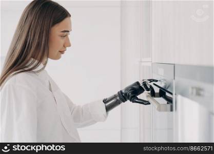 Disabled girl presses button on oven by finger of bionic prosthetic arm. Woman with artificial hand cooking preparing dinner with modern kitchen appliance. High tech prosthesis advertising.. Disabled girl presses button on oven by finger of bionic prosthetic arm. High tech prosthesis ad