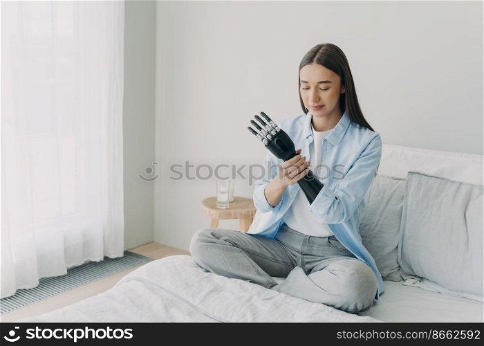 Disabled girl puts together her bionic prosthetic arm, adjusting grasp, position of artificial limb, sitting on bed at home. Lifestyle of people with disabilities. Advertising of high tech prosthesis.. Disabled girl puts together bionic prosthetic arm, adjusts position of artificial limb at home