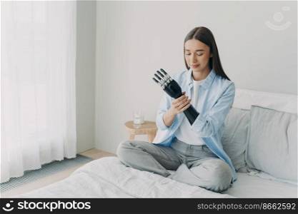 Disabled girl puts together her bionic prosthetic arm, adjusting grasp, position of artificial limb, sitting on bed at home. Lifestyle of people with disabilities. Advertising of high tech prosthesis.. Disabled girl puts together bionic prosthetic arm, adjusts position of artificial limb at home