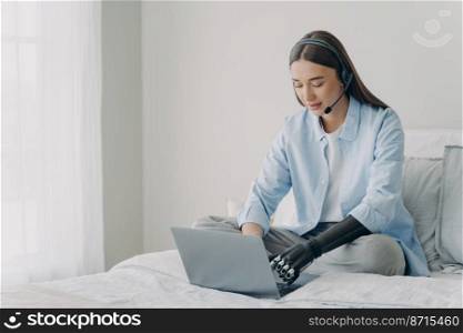 Disabled girl student wearing headphones, learning online at laptop at home. Young woman in headset typing using bionic prosthetic arm, sitting on bed. Lifestyle of people with disabilities.. Disabled girl in headset typing on laptop using bionic prosthetic arm, sitting on bed at home