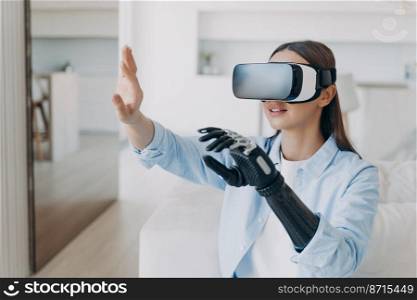 Disabled girl wearing virtual reality glasses with bionic prosthetic arm gets rehabilitation. Woman in vr goggles raising robotic hand interacting with objects in cyberspace. Medical high tech concept. Disabled girl wearing virtual reality glasses with bionic prosthetic arm gets rehabilitation