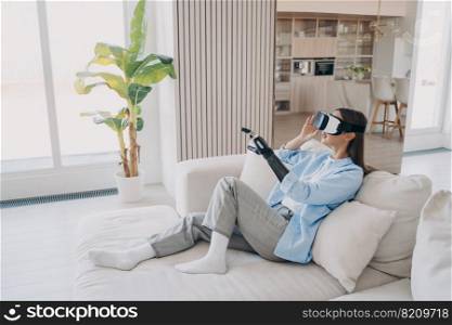 Disabled girl with artificial limb wearing virtual reality glasses learning to use bionic prosthetic arm during rehabilitation after limb loss, sitting on sofa in cozy living room. Medical high tech.. Disabled girl with in virtual reality glasses learning to use prosthetic arm in cozy living room