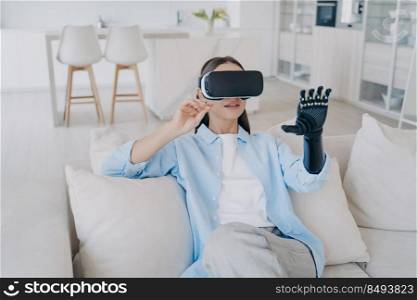 Disabled girl with cyber arm in vr glasses. Handicapped person gets rehabilitation at home. Young attractive european woman is sitting in living room on couch. Healthcare innovation technology.. Disabled girl with cyber arm in vr glasses. Handicapped person gets rehabilitation at home.