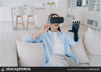 Disabled girl with cyber arm in vr glasses. Handicapped person gets rehabilitation at home. Young attractive european woman is sitting in living room on couch. Healthcare innovation technology.. Disabled girl with cyber arm in vr glasses. Handicapped person gets rehabilitation at home.